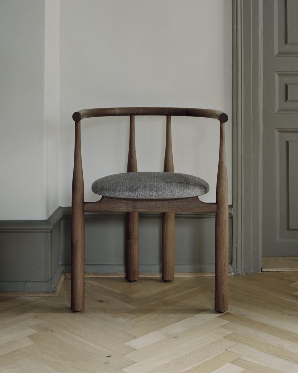 En træstol med et buet ryglæn og et gråt polstret sæde, placeret på et sildebensparketgulv i et rum med grå vægge og indviklet støbning, der eksemplificerer raffineret interiørdesign.