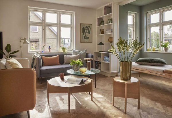 En lys, rummelig stue med moderne interiør. Møblerne omfatter en sofa, lænestole og et sofabord. Store vinduespartier giver naturligt lys, og værelset har et sildebensgulv med planter og kunst.