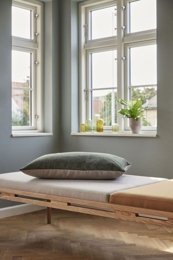 Et fredfyldt værelse med store vinduer, hvide rammer og en plysgrøn pude på en træbænk fra TomorrowsDesign. Vinduerne afslører en fjern udsigt udefra, der omfatter en rolig atmosfære.