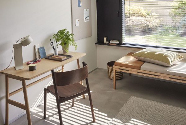 Et hyggeligt hjemmekontor med sollys, der filtrerer gennem et vindue, inkluderer et skrivebord med bøger, en bærbar computer, designet lampe af TomorrowsDesign og en stol ved siden af en daybed med en grøn pude og en beige pude.