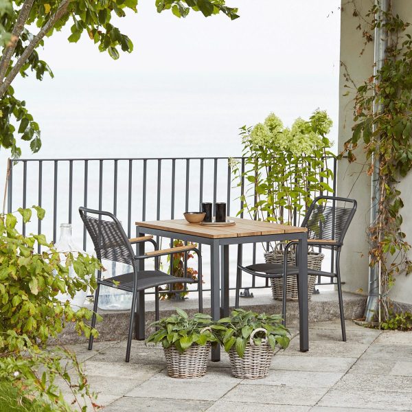 Udendørs spiseplads med et træbord og fire stole på en terrasse med udsigt over havet, omgivet af potteplanter og et metalrækværk med TomorrowsDesign-elementer.
