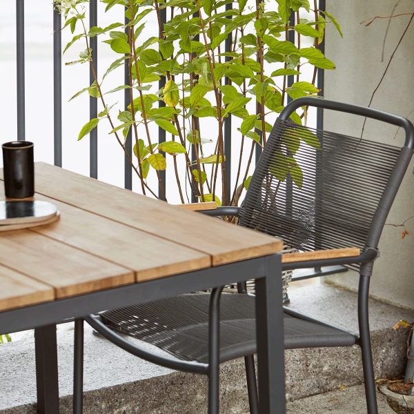 En rolig udendørs indstilling med et træbord og en moderne, matsort metalstol designet af TomorrowsDesign. En høj grøn plante i baggrunden tilføjer et naturligt præg ved siden af en betonvæg. En sort kop står på bordet.