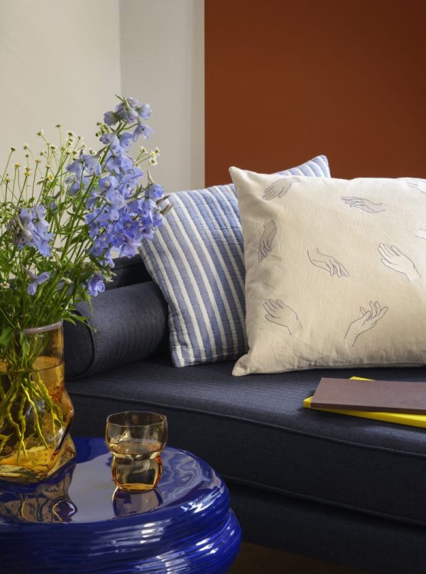 Et hyggehjørne med en BROSTE COPENHAGEN - 'Wind' Sofa 2-Personers prydet med stribede og mønstrede hynder. et blåt keramikbord rummer et glas whisky, en notesbog og en vase med blå blomster. varm og indbydende indretning.