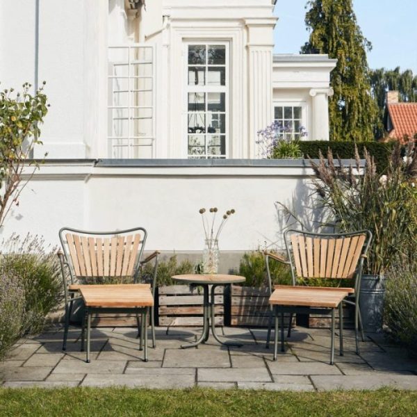 To træ- og metalstole fra TomorrowsDesign står over for hinanden på tværs af et lille rundt bord på en flisebelagt terrasse, med frodigt grønt og en del af en hvid bygning synlig i baggrunden.