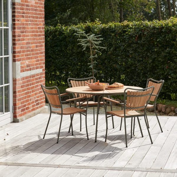 Udendørs terrasse med et spisebord af træ og fire stole på et dæk ved siden af en murstensbygning, omgivet af frodige grønne områder. En baguette og kande er placeret på bordet under et lille pottetræ, der viser elegante designmøbler.