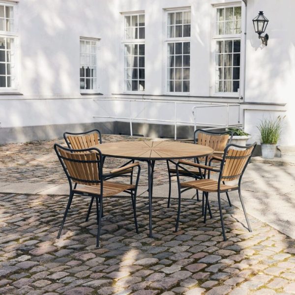 Et hyggeligt udendørs opholdsområde med et rundt træbord og fire matchende stole på en brostensbelagt gårdhave, flankeret af en lys hvid bygning med store vinduer og en sort lanterne med skandinavisk designmøbler.