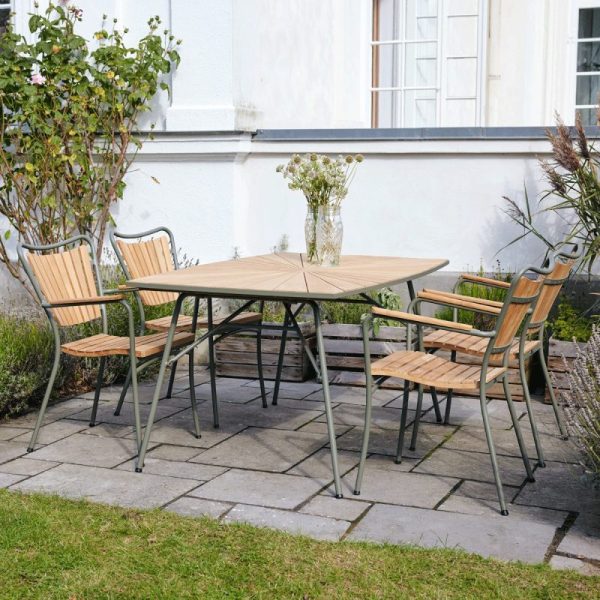 Et terrassesæt med et træbord og fire stole, placeret på en stenterrasse ved siden af et hus med hvide vægge med lidt grønt omkring, viser TomorrowsDesigns karakteristiske design.