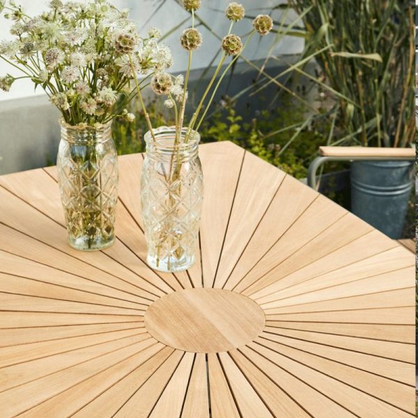 Rundt træbord med et radialt design med en centreret cirkulær planke, ledsaget af to glasvaser med vilde blomster på en terrasse med synlige grønne planter i baggrunden. Dette eksempel på TomorrowsDesign bringer moderne æstetik til klassiske former.