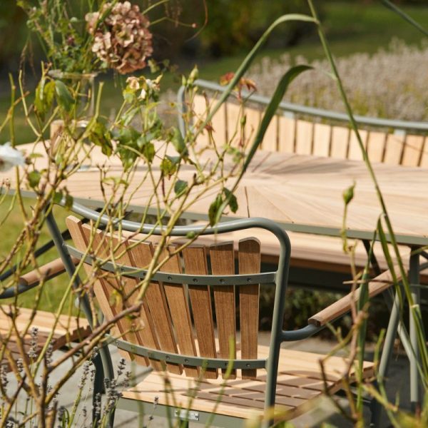 En udendørs stol i træ med armlæn i gråt metal og et moderne design sidder ved siden af et træbord, omgivet af frodigt grønt og bløde blomster i rolige havemiljøer.