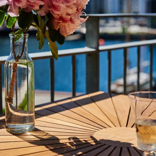 Et bord med en glasflaske med en gren og lyserøde blomster, ved siden af et glas vand, med udsigt over en solrig havn med både. Designet understreger enkelhed og elegance i sit arrangement.