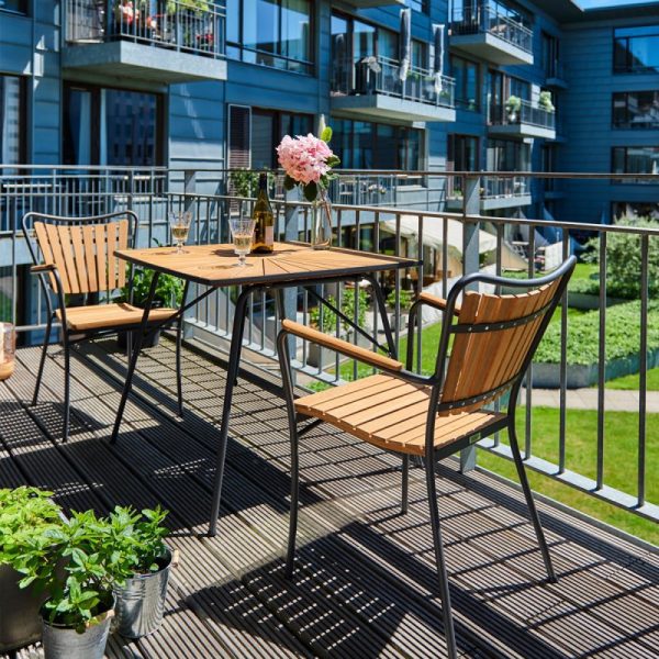 Udendørs balkon i en moderne lejlighed med designmøbler inklusiv et træbord med to stole og en vase med lyserøde blomster. Frodigt grønt omgiver området og forstærker den hyggelige, indbydende stemning.