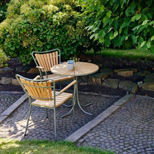En fredelig have med et lille cirkulært bord og to stole, designet med sans for detaljer, omgivet af frodige grønne planter og brostensbelagte fortov. En enkelt kop hviler på bordet.