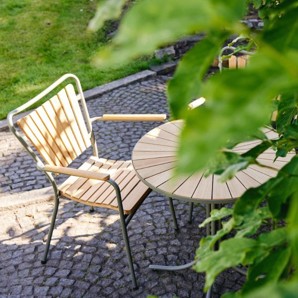Havemøbler af træ designet af TomorrowsDesign, bestående af et rundt bord og en stol, sat på en brostensbelagt terrasse omgivet af frodigt grønt løv i skarpt sollys.