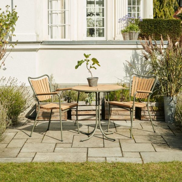 En terrasse med et rundt træbord og fire designmøbler stole sat på belægningssten, prydet af en potteplante, omgivet af frodige planter under sollys nær en hvid husmur.