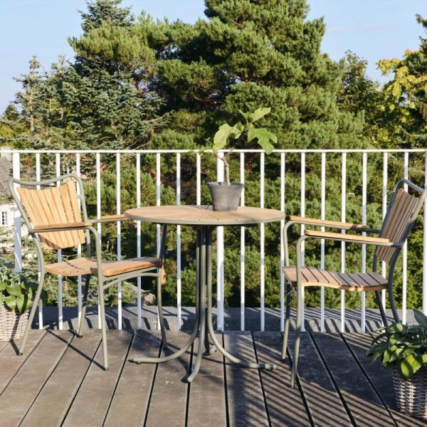 En lille udendørs terrasseramme med to træstole og et rundt bord fra TomorrowsDesign, placeret på en terrasse omgivet af frodigt grønt og træer under en klar himmel.