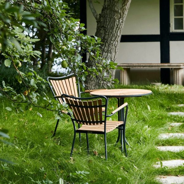 En træstol og et rundt bord sat på en frodig grøn græsplæne ved siden af en brostensbelagt sti med et bindingsværkshus og grønne træer i baggrunden, der afspejler TomorrowsDesign-æstetik.