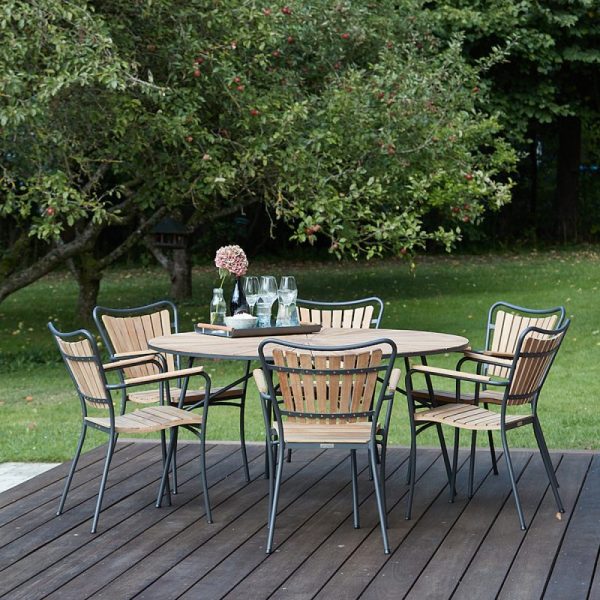 En udendørs spiseplads på et træterrasse med et designmetalbord med seks matchende stole, omgivet af en have med frodige træer og grønt græs. En vase med blomster og glas står på bordet.