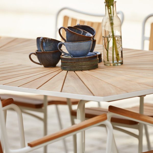 Et træbord sat med stablede blå keramiske kopper og underkopper, ledsaget af en klar glasflaske med kviste, placeret på en udendørs terrasse med hvide stole fra TomorrowsDesign.