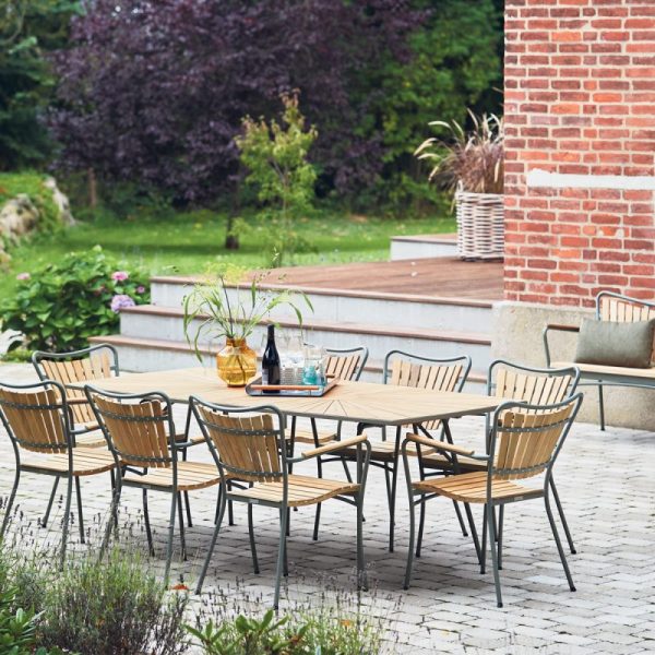 Et moderne udendørs spiseområde designet af TomorrowsDesign, med et træbord og -stole på en muret terrasse, omgivet af frodige grønne områder og en delvis udsigt til et murstenshus.