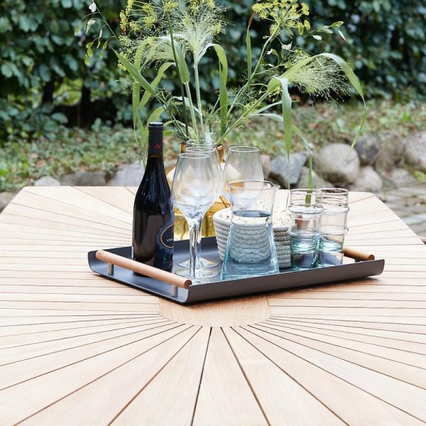 En bakke med en flaske vin, glas og krukker pakket ind i sejlgarn står på et cirkulært træbord udendørs, designet til en perfekt blanding af funktion og stil, omgivet af frodige grønne områder.