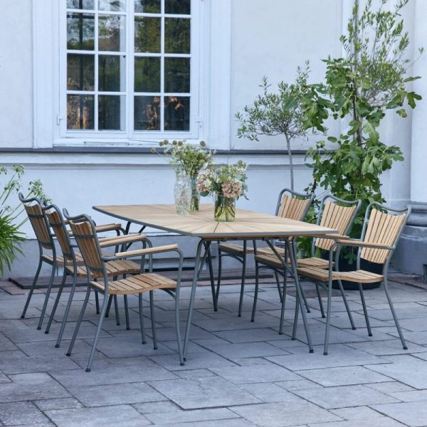 Et gårdhave spisebordssæt bestående af et træbord og seks stole med metalstel og trælameller, arrangeret på en stenterrasse. Potteplanter og afskårne blomster tilføjer et naturligt præg til scenen, hvilket afspejler etos i TomorrowsDesign.