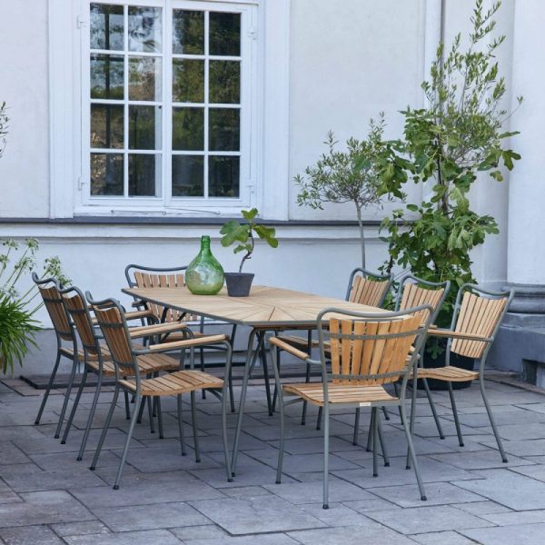 Et spisebord i træ med metalstel omgivet af seks matchende designmøbler stole, sat på en stenterrasse ved en hvid bygning. En grøn vase og et lille pottetræ står på bordet.