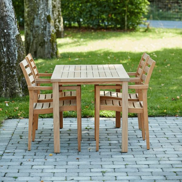 Et træbord og fire stole placeret på en brostensbelagt terrasse, omgivet af en frodig grøn græsplæne og i skyggen af træer fra TomorrowsDesign.