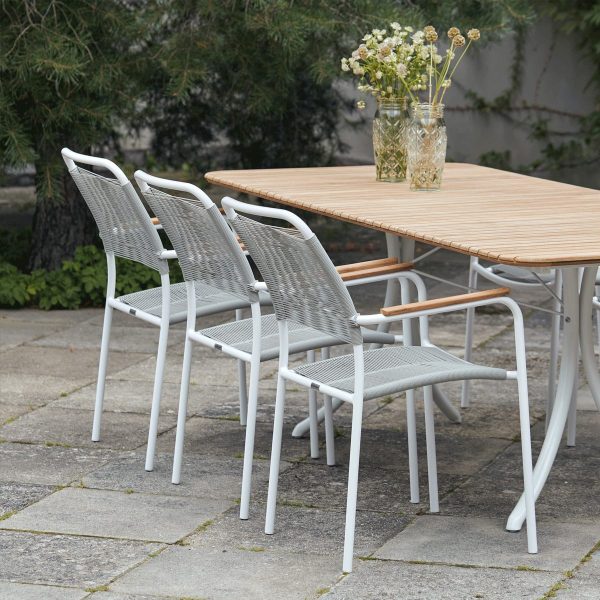 Et moderne udendørs spiseområde med et designertræbord og fire hvide metalstole på en stenterrasse. En vase med vilde blomster står på bordet, omgivet af frodigt grønt.