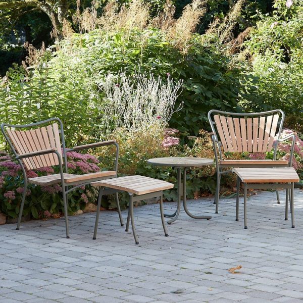 Et sæt havemøbler i træ bestående af to stole og en bænk, arrangeret på en cirkulær stenterrasse, omgivet af frodige grønne planter og blomstrende planter i en TomorrowsDesign-inspireret have.