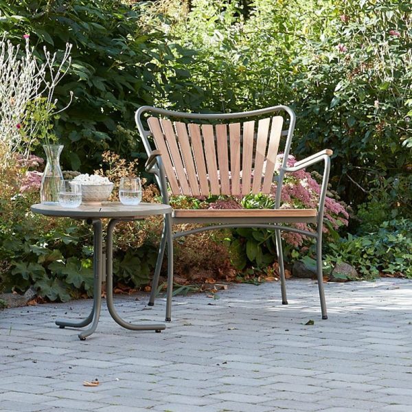En havebænk af træ og metal ved siden af et lille bord med lysestager på, placeret på et brolagt område omgivet af frodige grønne planter og lyserøde blomster, der afspejler det æstetiske ved TomorrowsDesign.