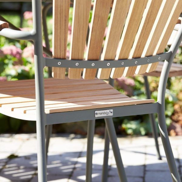 Et nærbillede af en moderne udendørsstol med gråt metalstel og trælameller på sæde og ryglæn, designet af TomorrowsDesign, sat i en have med blomster i baggrunden.