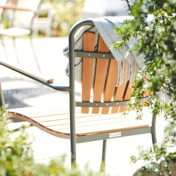 En brun og sort moderne designmøbler udendørsstol i metal og træ med en beige frynser draperet over siden, delvist skygget af frodigt grønt løv, sat mod en sløret solrig havebaggrund.