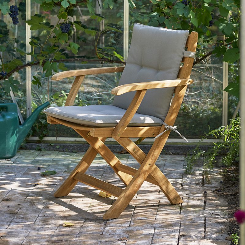 En træfoldestol med en grå hynde på en muret terrasse, omgivet af frodige grønne planter og sollys, der filtrerer gennem bladene, repræsenterer et elegant stykke møbler, der er perfekt til ethvert udendørsrum.
