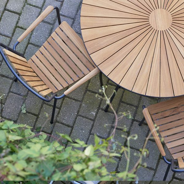 Set ovenfra og ned af et rundt træbord med solstrålemønster og to matchende stole på en brostensbelagt terrasse, omgivet af grønne planter og forstærket af designmøbler-æstetik.