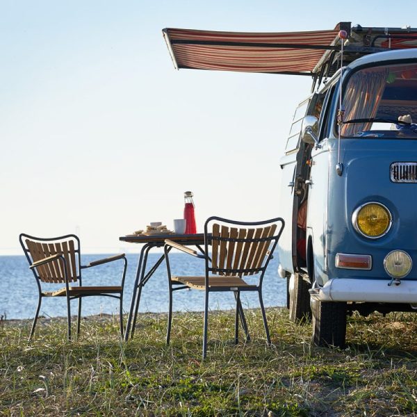 En vintage blå varevogn parkeret ved havet med en stribet markise strakt ud over et lille bord og to stole, designet til et måltid, med udsigt over et roligt hav under en klar blå himmel.