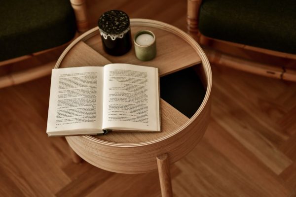 En åben bog ligger på et rundt træbord ved siden af et tændt stearinlys og en lille kop, i et hyggeligt rum med to designmøbler stole og et varmt tonet gulv.