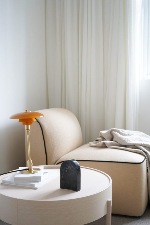 En hyggelig læsekrog med en beige sofa med et blødt tæppe, et rundt sidebord med bøger og et unikt sort stenur, alt sammen suppleret af designmøbler. Placeret ved siden af et vindue draperet med hvide gardiner og en orange lampe.