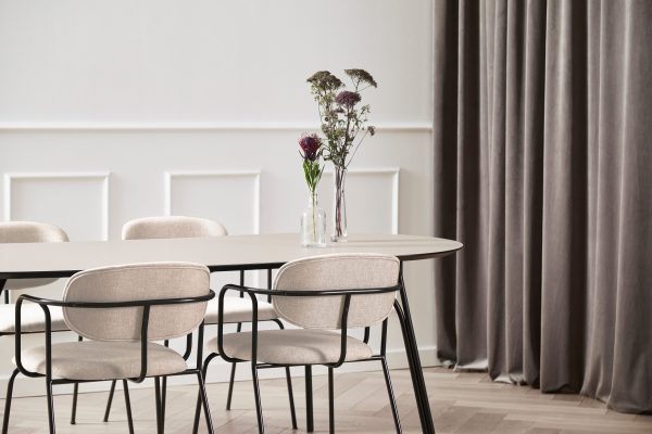 En moderne spisestue med et ovalt træbord med fire beige stole, sat mod en hvid panelvæg og lange grå gardiner. En vase med lilla og hvide blomster pryder bordet og er et eksempel på et omhyggeligt interiørdesign.