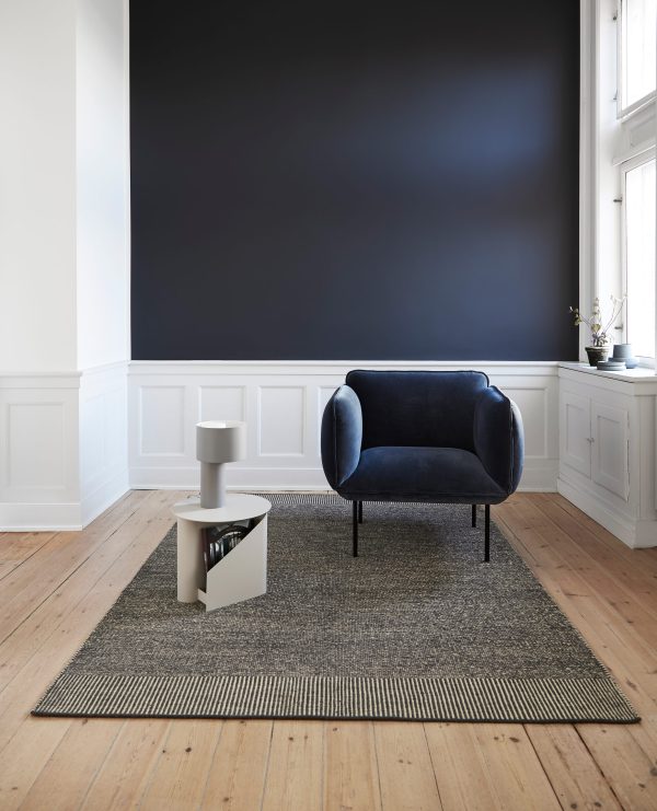 En moderne stue med et WOUD - Nakki Lænestol centreret på et stribet tæppe, med et hvidt sidebord og en plante i baggrunden mod en mørk væg og hvidt waincoting.