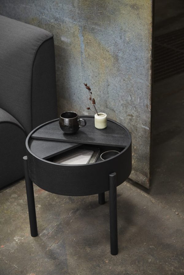 Et lille, cirkulært, sort sidebord med en forsænket plade med en kop, en lille kande og en bog fra TomorrowsDesign, ved siden af en grå lænestol mod en gammel metalvæg.