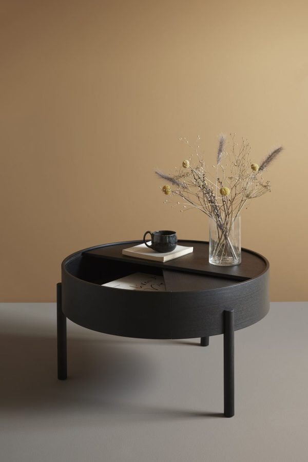 Et moderne design, mørkt cirkulært sofabord på en solid beige baggrund. Bordet rummer en bog, en sort kaffekop og en klar vase fyldt med tørrede blomster.
