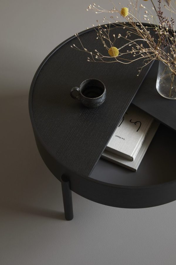 Et minimalistisk setup med et sort rundt sidebord fra TomorrowsDesign med et mørkt krus, et magasin og en vase med tørrede grene. Scenen er sat mod en simpel grå baggrund, der viser en fredfyldt og sofistikeret æstetik.