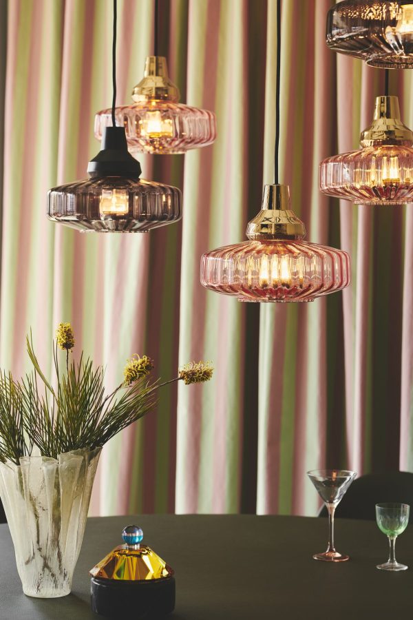 Moderne hængelamper med forskellige farvede glasskærme oplyser et stilfuldt interiør med stribede gardiner, et blomsterarrangement og dekorative glasvarer på en overflade nedenunder, hvilket afspejler principperne i TomorrowsDesign.