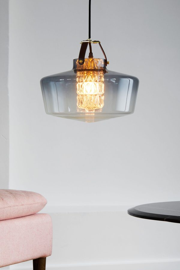A DESIGN BY US - Addicted To Us Lampe med et røgfyldt glasdæksel og indviklede guldindvendige armaturer, hængende over en sløret lyserød stol og et sort bord mod en grå baggrund.