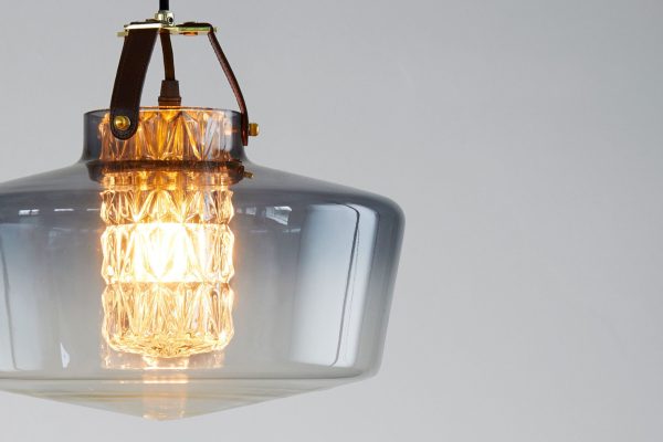 En moderne pendel med en gennemsigtig glasskærm og en lys, indviklet designet glødetrådspære, der hænger fra en gylden armatur mod en lysegrå baggrund, som DESIGN BY US - Addicted To Us Lampe.