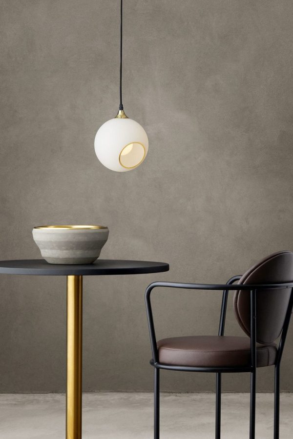 Et minimalistisk interiør med DESIGN BY US - Ballroom Lampe, der hænger over et bord med en guldbund og en mørk bordplade, ved siden af en mørkebrun stol, mod en tekstureret grå væg.