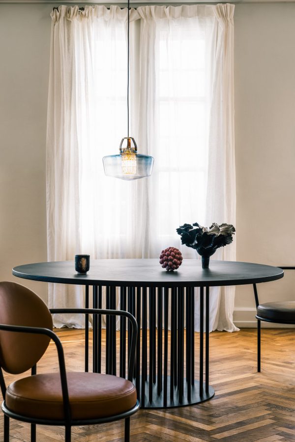 Et veloplyst spiseområde med et rundt sort bord med to brune læderstole, en skål med vindruer og sorte blomster på bordet under en unik pendel (DESIGN BY US - Addicted To Us Lampe) ved siden af rene gardiner.
