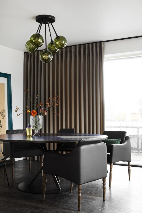Moderne spisestuedesign med et sort rundt bord med stole, olivengrønne glaslysekroner ovenover og gulvlange brune gardiner. En vase med blomster tilføjer et strejf af farve.