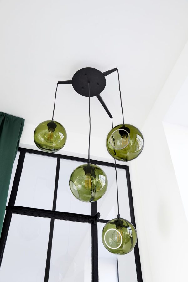 A DESIGN BY US - Ballroom Lampe XL med fire grønne glasskærme, der hænger i forskellige højder fra en sort metalramme, monteret på et hvidt loft over en sort vinduesramme.