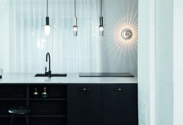 En moderne køkkenbord med en dyb sort vask med en høj vandhane, flankeret af cylindriske pendellamper og en DESIGN BY US - New Wave Optic Væglampe, der støber et radialt mønster.